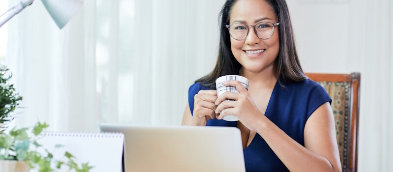 A Filipino virtual assistant with glasses sits at her desk, holding a coffee mug while focused on work.