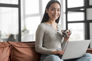 A woman sitting on a sofa holding a mug and using a laptop offering virtual assistants services.